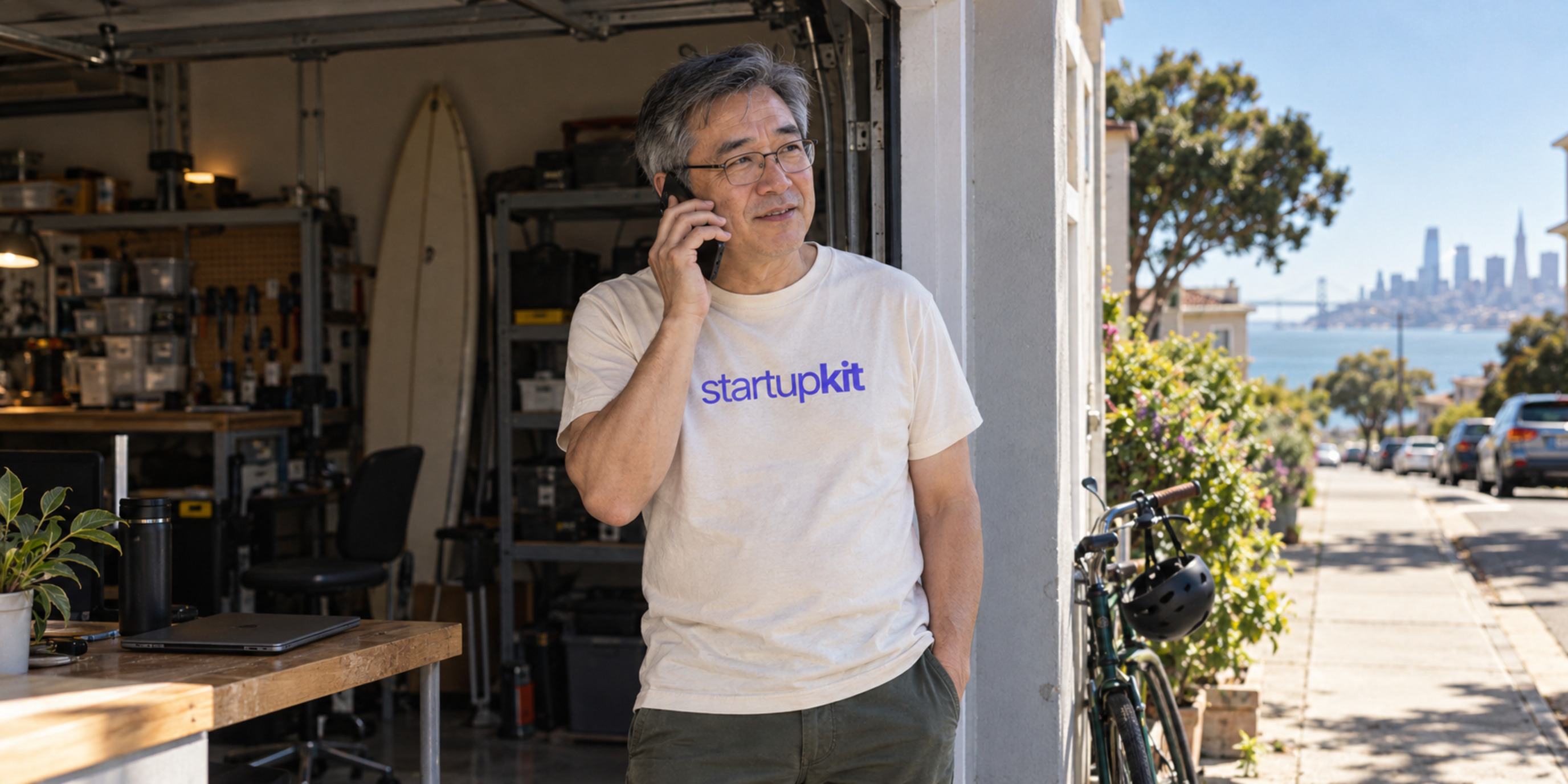 An older East Asian man wearing a StartupKit shirt talks on the phone beside a closed laptop at a sunny San Francisco garage doorway.