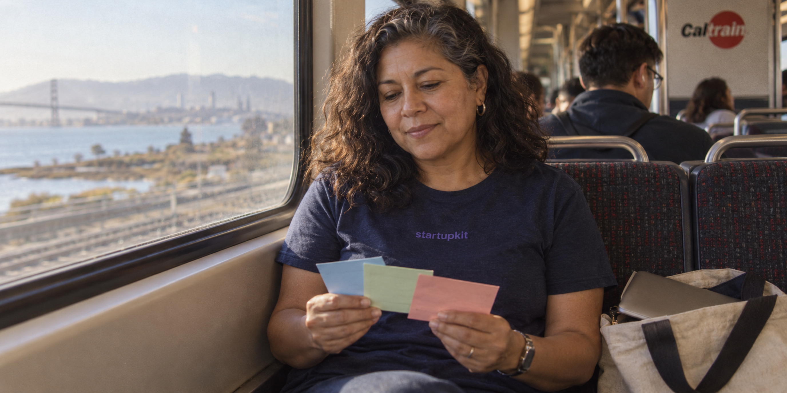 A Latina talent lead in her 50s reviews three blank cards beside a closed laptop on Caltrain, wearing a startupkit wordmark