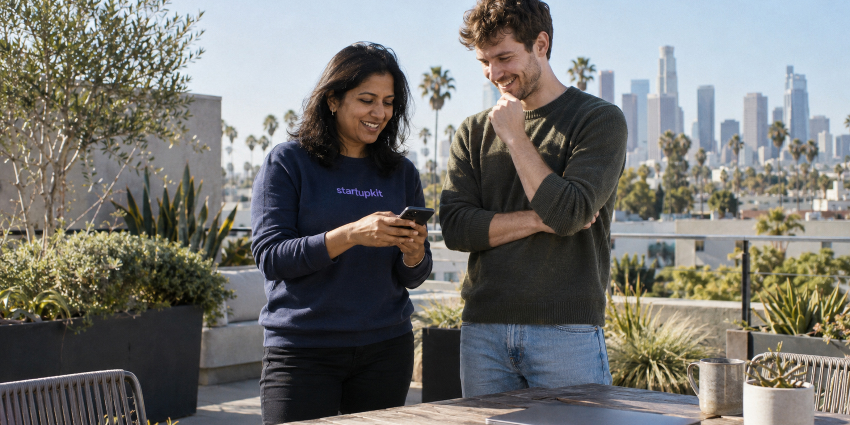 A South Asian woman in a navy startupkit sweatshirt and a male colleague check a phone beside a closed laptop on a sunny Los Angeles rooftop.