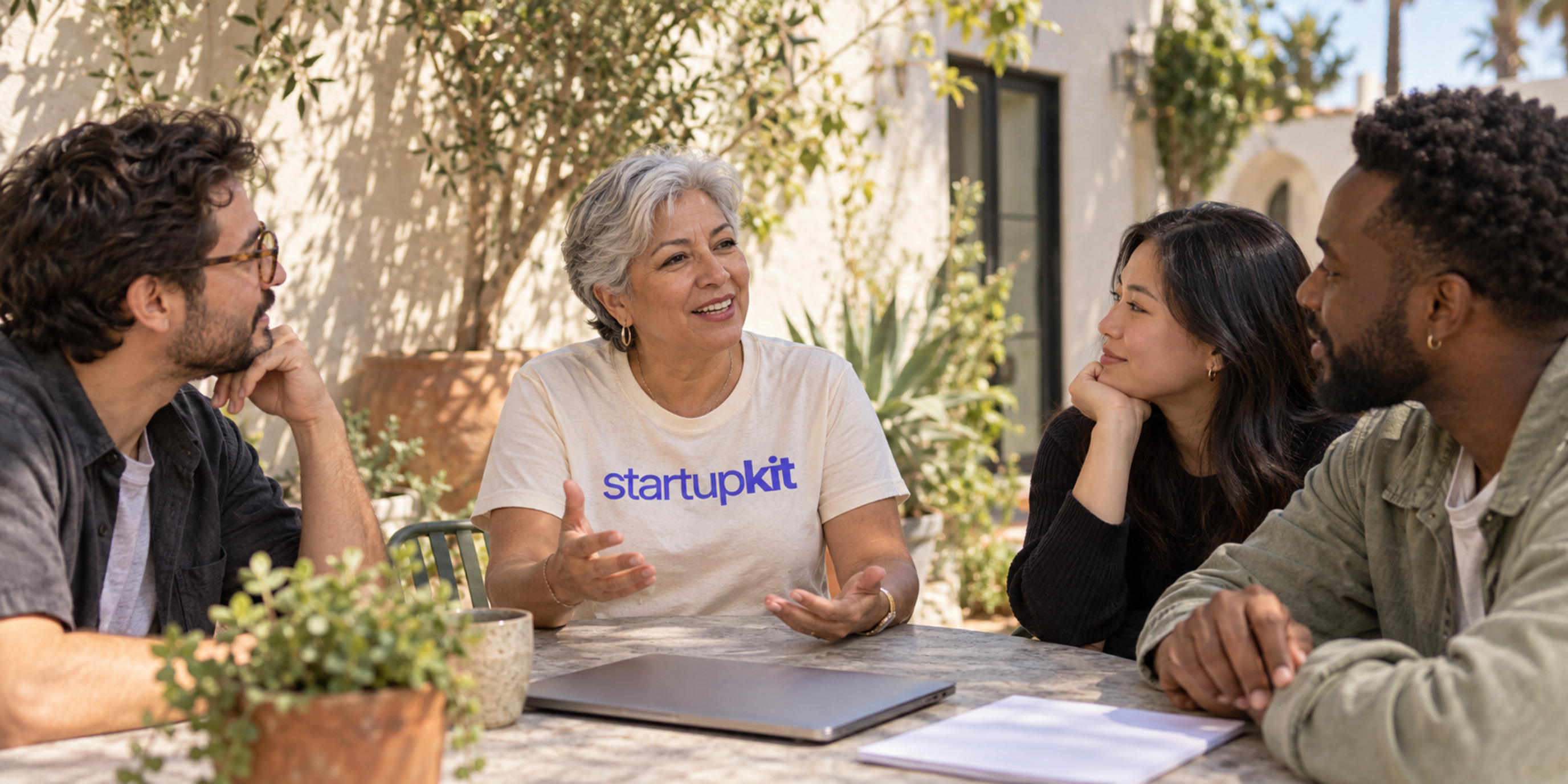 Four hiring colleagues talking in a sunlit Los Angeles courtyard, with an older Latina team lead in a cream startupkit shirt, a closed laptop, and face-down notes.