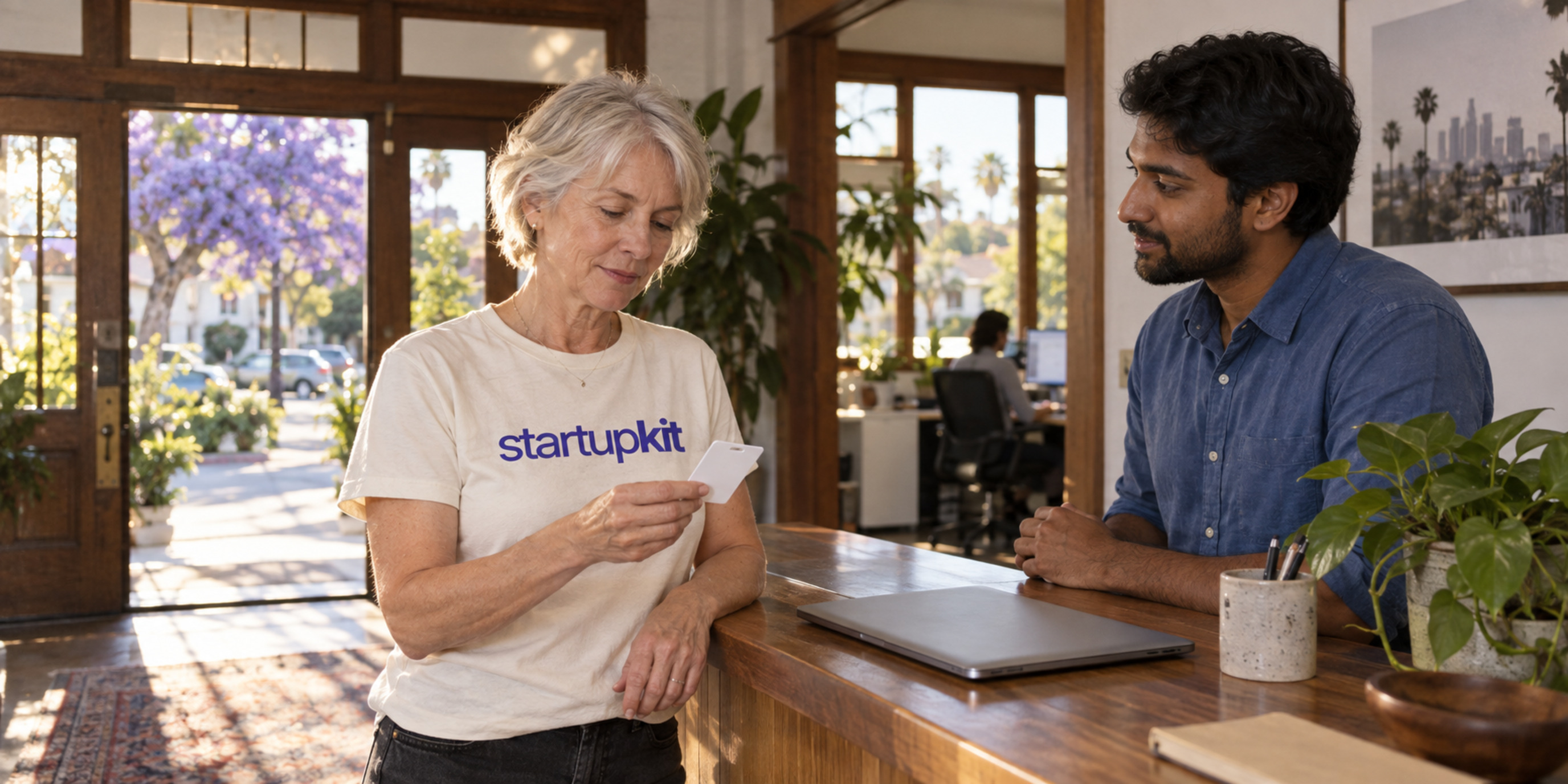 An older silver-haired woman holds a blank keycard at a Los Angeles Craftsman reception beside a younger male colleague and a closed laptop